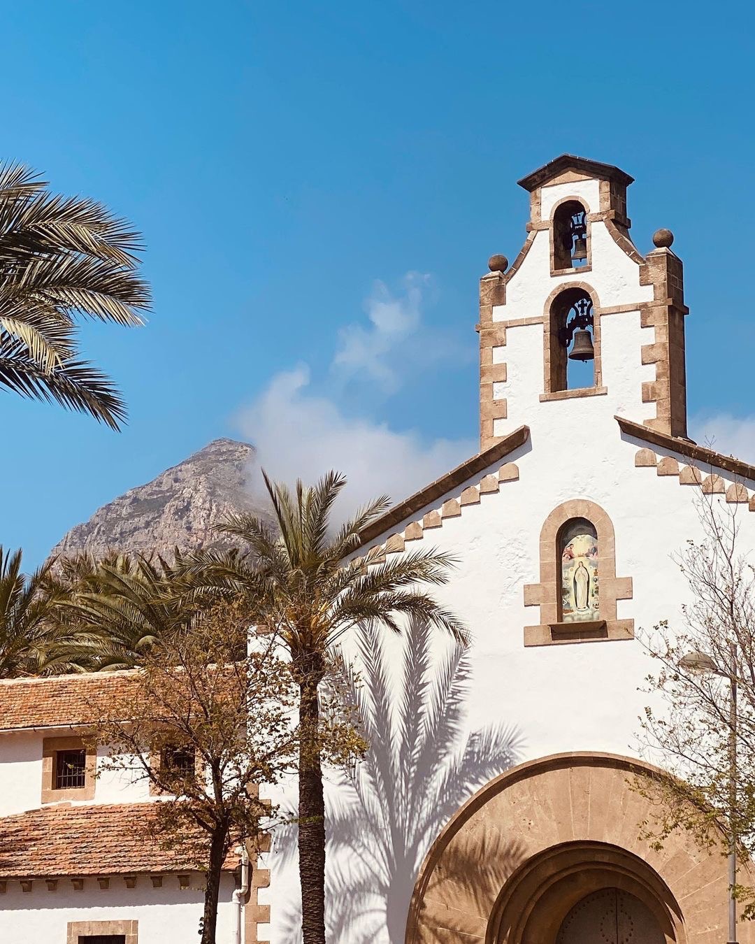 Iglesia blanca con campanario y palmeras bajo el Montgó